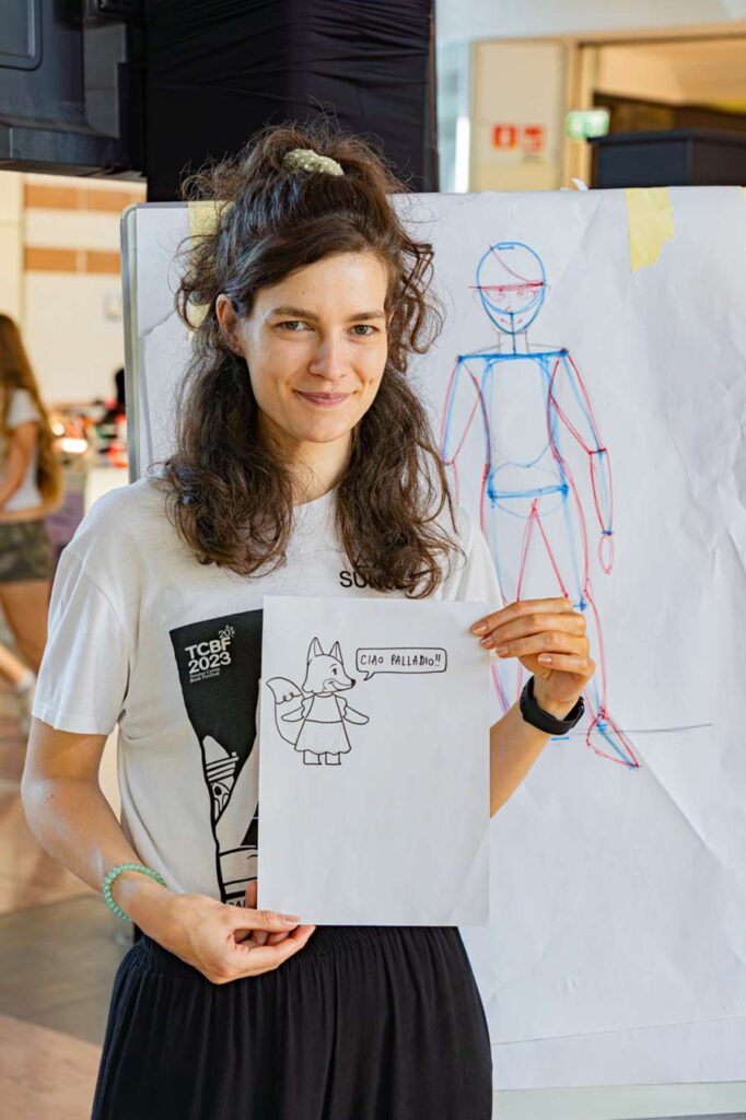 Una donna in piedi in piano americano sorride tenendo in mano un foglio dove si vede disegnata una volpe illustrata, sullo sfondo una lavagna mobile con fogli e disegnato uno scheletrino